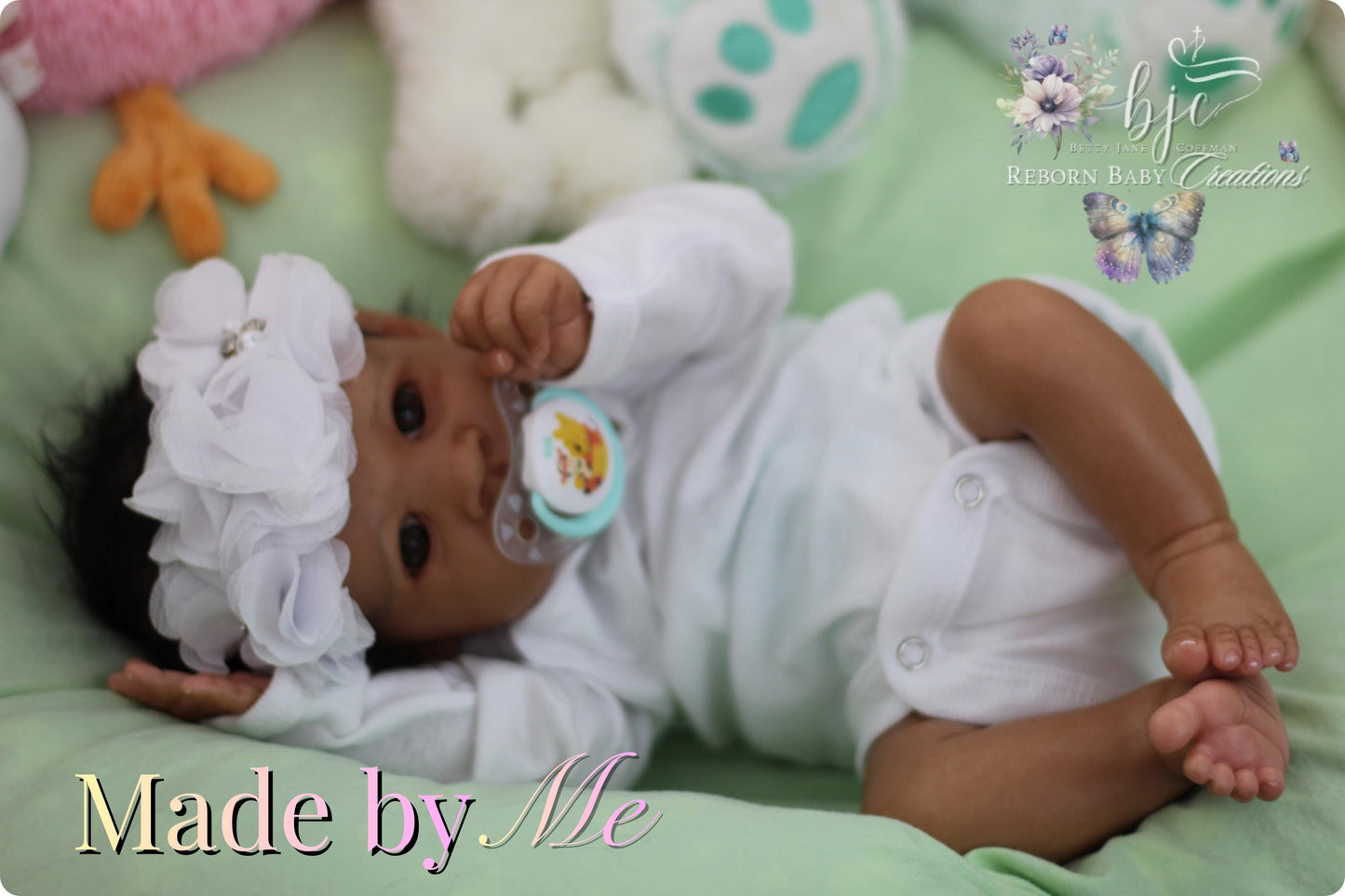 A handmade baby doll lying on a green blanket, wearing a white outfit and a white headband with a flower design.