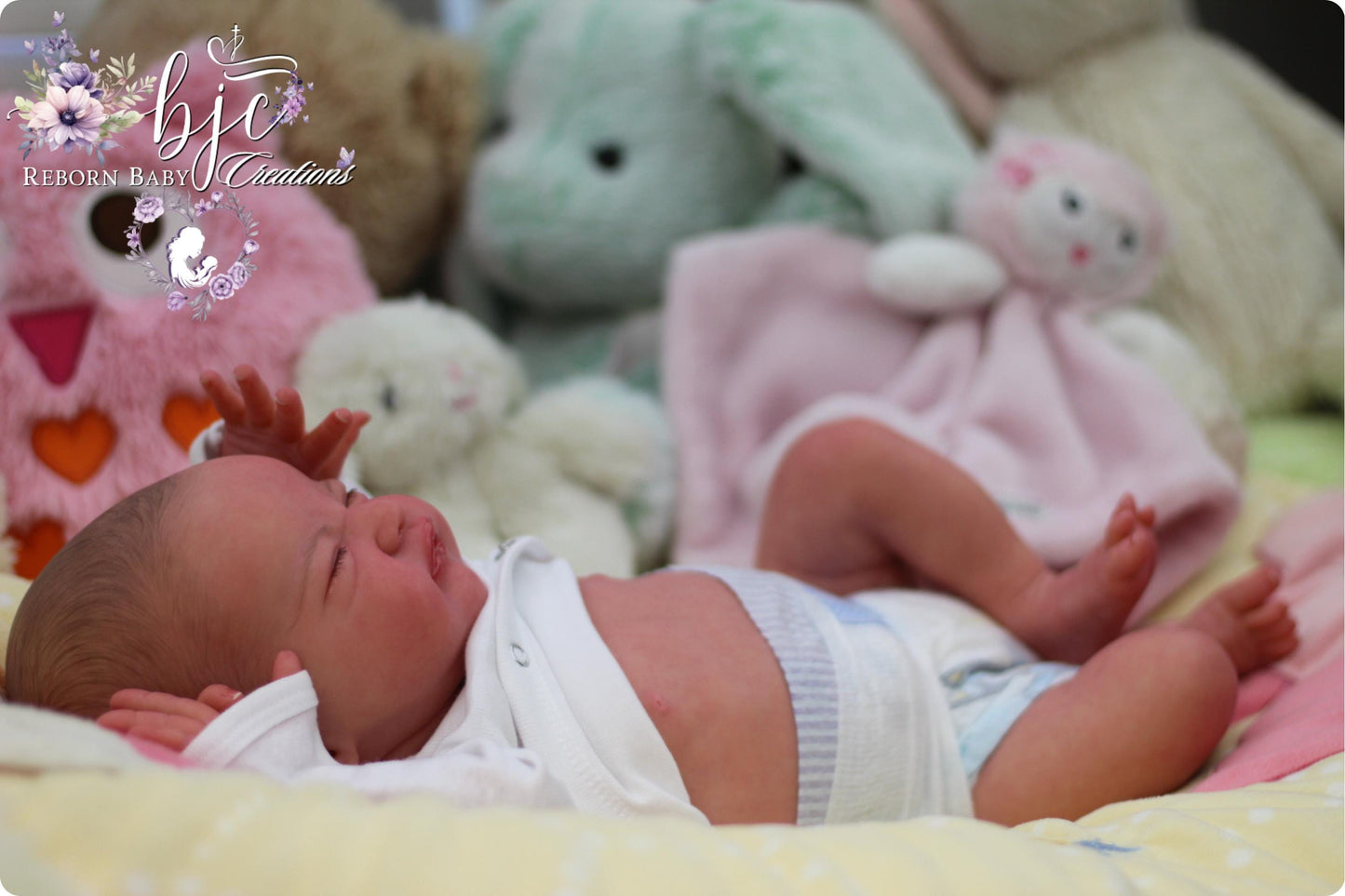 A newborn baby lies on a bed surrounded by various stuffed animals, including a pink bunny and a green bunny.